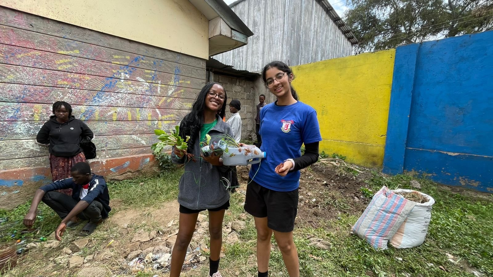 Two students holding a cut bottle planted with greens, with others working on the ground behind them