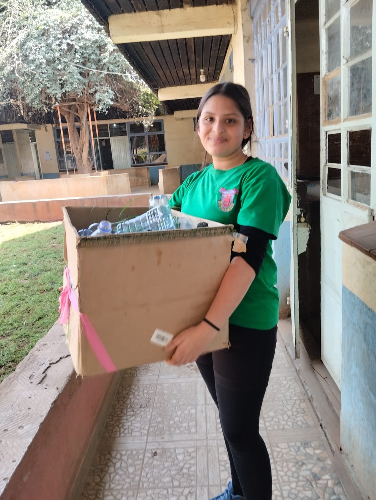 A student carrying a cardboard box of collected plastic bottles along a school corridor